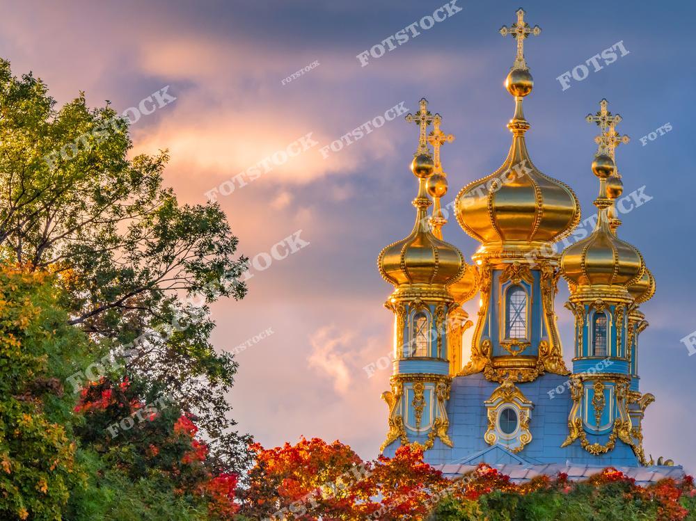 На фотографии показаны золотые купола Екатерининского дворца в Царском Селе, которые сияют на фоне облачного вечернего неба. Архитектура дворца в стиле русского барокко и осенние деревья с красно-золотыми листьями создают гармоничный и выразительный пейзаж. Эти купола являются символом Екатерининского дворца, привлекая внимание туристов своим великолепием. Свет заката добавляет фотографии особую теплоту и романтику, подчеркивая величие и красоту исторического памятника.
